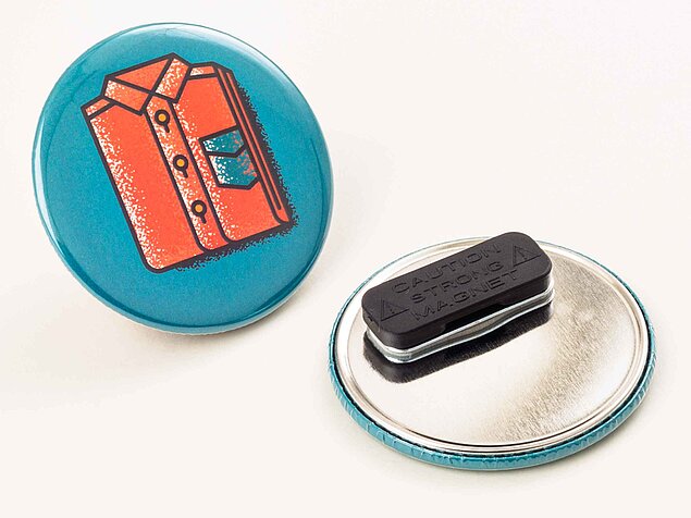 Zwei runde Kleidungsmagneten; eines zeigt eine orangefarbene Hemddarstellung auf blauem Hintergrund, das andere zeigt die metallische Unterseite mit einem rechteckigen Magnetclip. Die Umgebung ist hell.