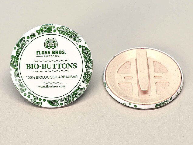 Zwei runde Buttons liegen auf einer Oberfläche. Ein Button zeigt folgenden Text: "FLOSS BROS. BUTTONS", "BIO-BUTTONS", "100% BIOLOGISCH ABBAUBAR", "www.flossbros.com". Der andere zeigt die Rückseite.