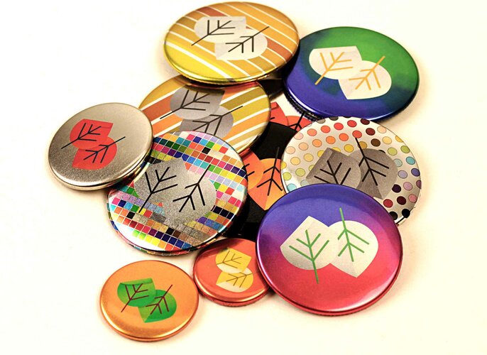 Mehrere bunte Ansteckbuttons liegen zusammen und zeigen stilisierte Blätter in verschiedenen Mustern und Farben, darunter Streifen, Punkte und ein Mosaikdesign, auf einem weißen Hintergrund.