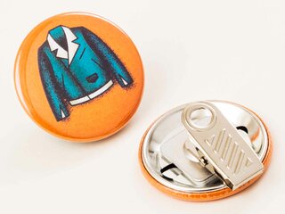 Ein runder Kroko-Clip-Ansteckbutton zeigt eine blaue Jacke auf orangem Hintergrund. Ein zweiter Clip-Button liegt daneben, Rückseite mit Metallclip sichtbar. Beide liegen auf einer hellen Oberfläche.