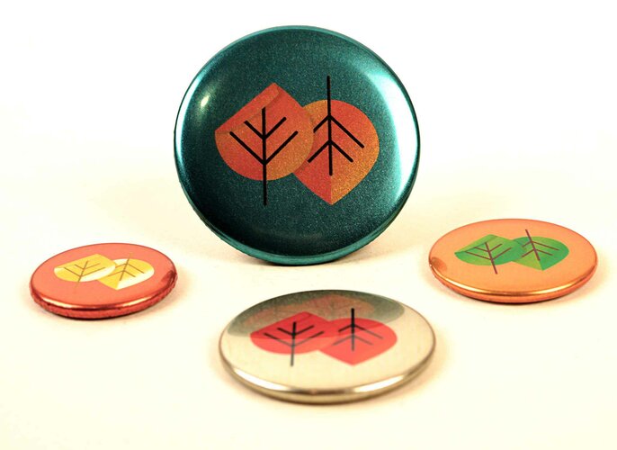 Vier runde Ansteckbuttons mit Foliendruck Veredlung mit stilisierten Blattmotiven in Herbstfarben liegen auf einer hellen Oberfläche. Der größte Button steht aufrecht, während die anderen flach liegen.