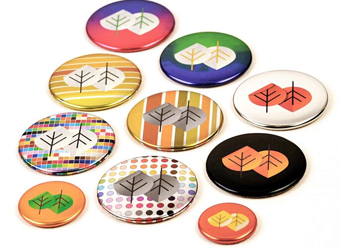 Verschiedene bunte Buttons veredelt mit Metallic-Chrome zeigen stilisierte Blätter in verschiedenen Farbkombinationen. Sie liegen auf einer weißen Oberfläche und variieren in Größe und Muster, einschließlich Streifen und Punkten.
